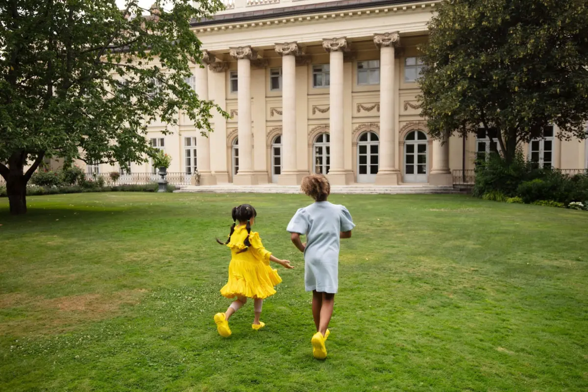 Two children in yellow and blue dresses run across a green lawn toward a beige, columned building.