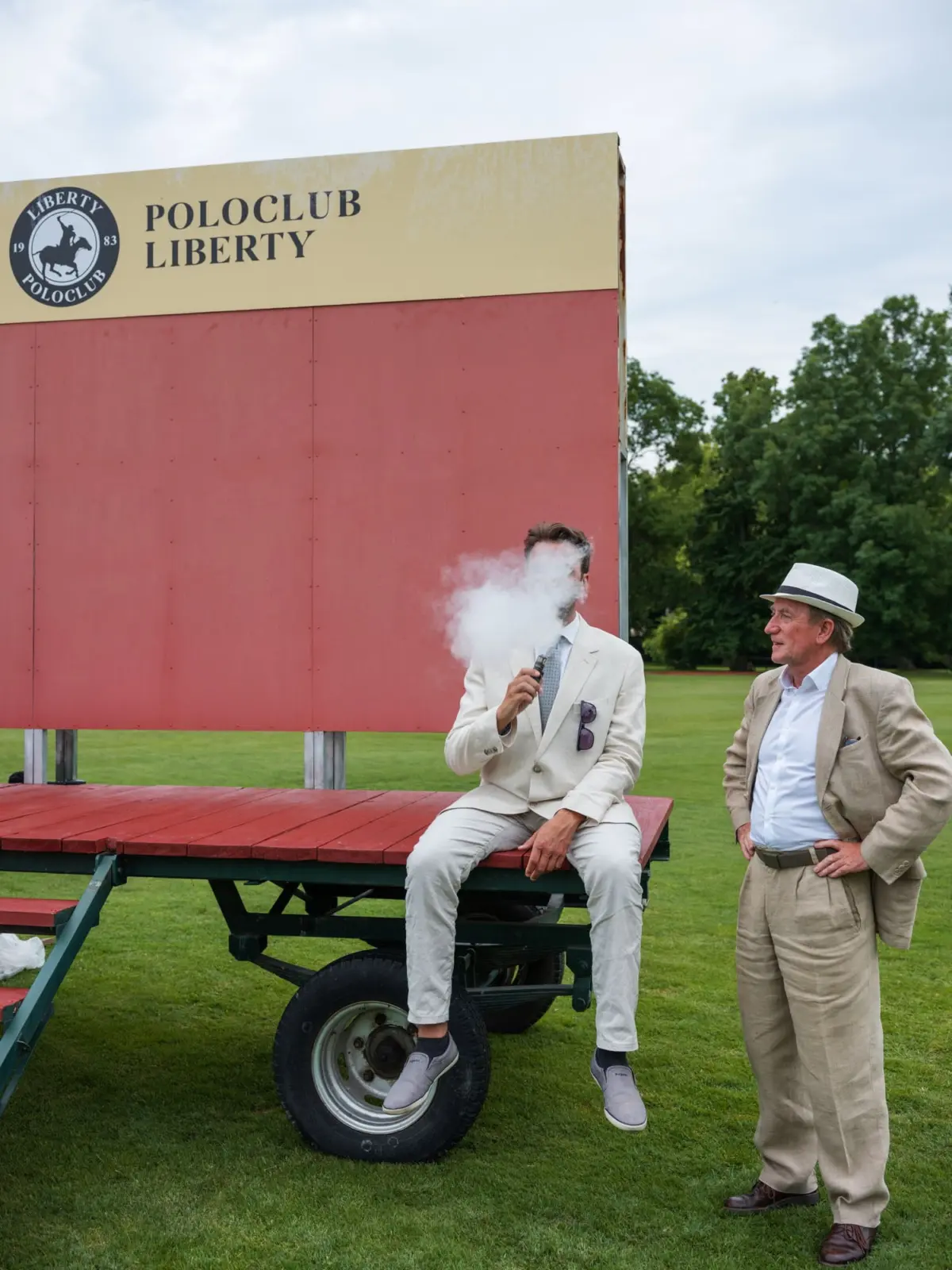 Two people in beige suits on a red platform at a polo club; one sits vaping, blowing vapor; a beige sign reads POLOCLUB LIBERTY behind them.