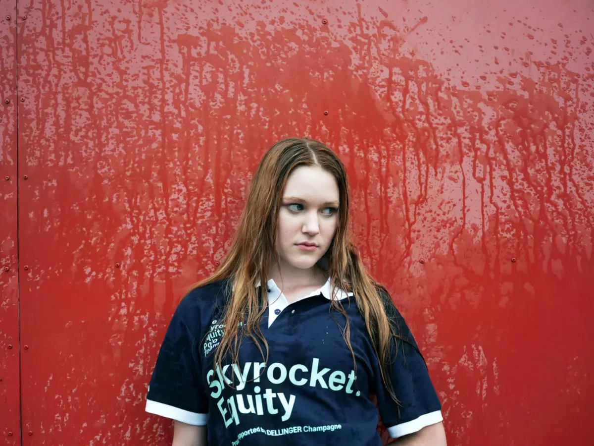 Person with long brown hair wearing a navy shirt stands in front of a red wall with dripping, peeling paint.