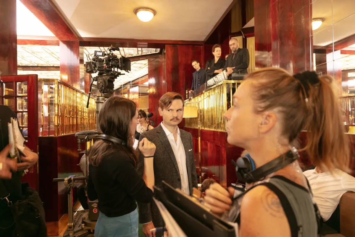 Film crew operates a cinema camera in a mahogany-paneled lobby with brass railings as suited actors converse on a balcony.