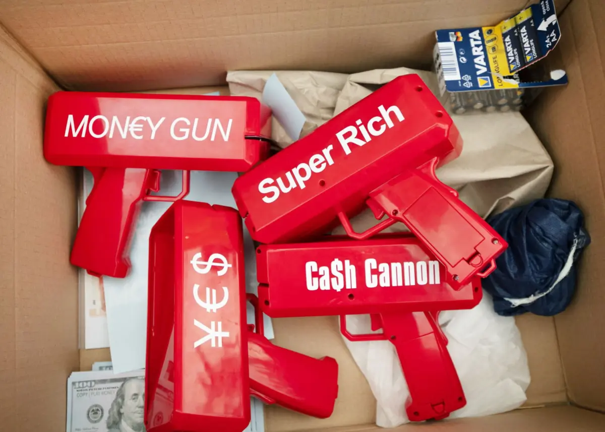 Red money guns labeled Money Gun, Super Rich, and Cash Cannon in a cardboard box with packing paper.