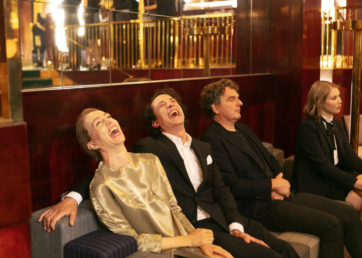 Four adults (two women, two men) in formal attire laugh while seated on a sofa in a gold-toned hotel lounge.