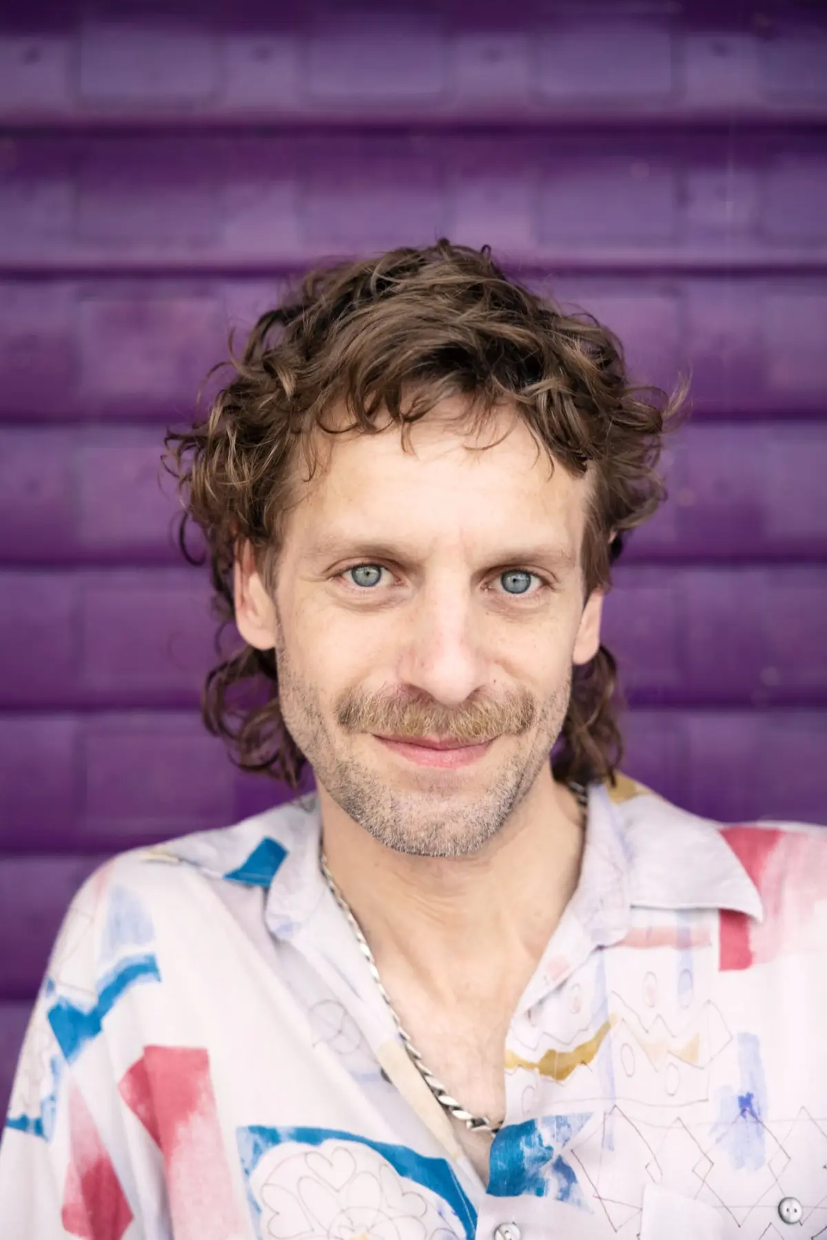 Adult person with wavy brown hair and blue eyes smiles at camera, wearing a colorful patterned shirt, in front of a purple wall.