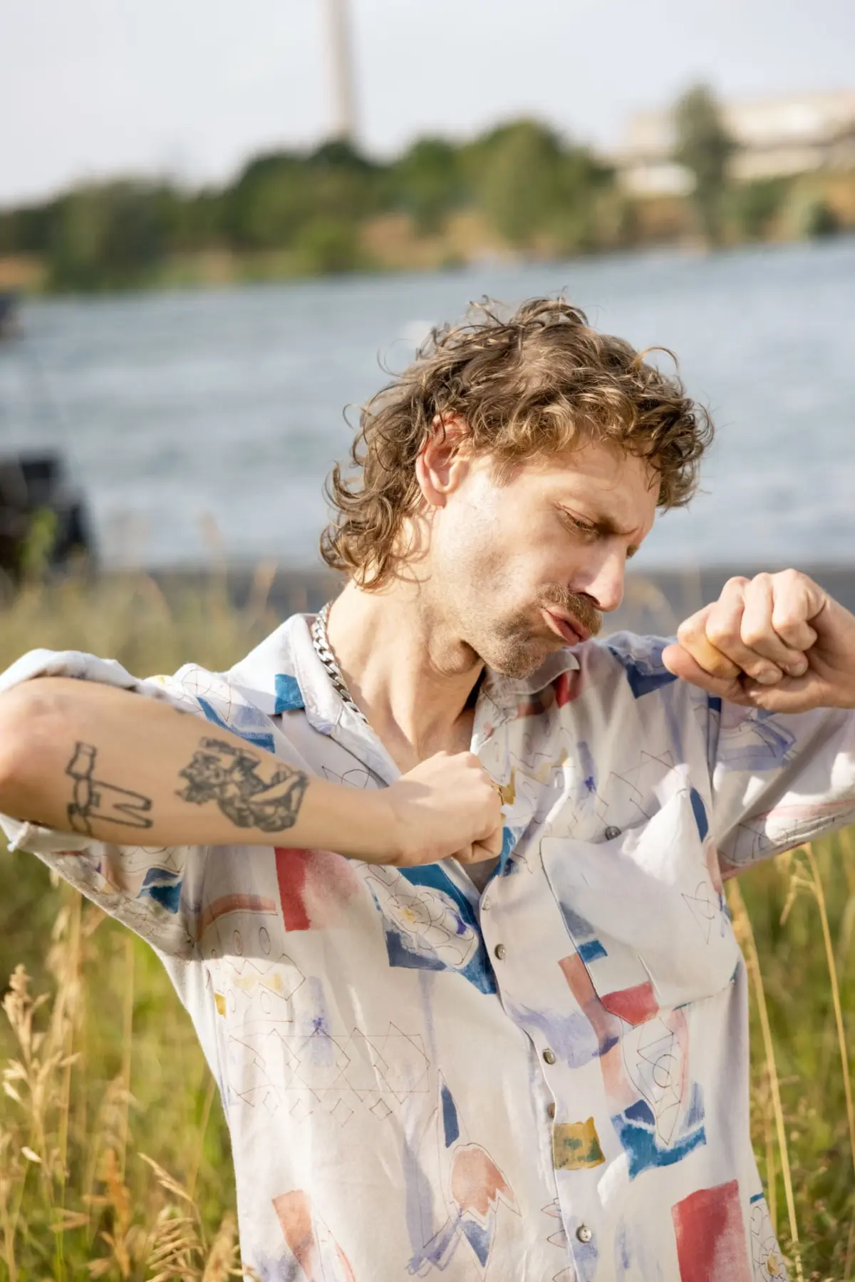 Adult man with wavy brown hair and arm tattoos, flexing his arms by a body of water, wearing a colorful abstract-pattern shirt; grasses in foreground.