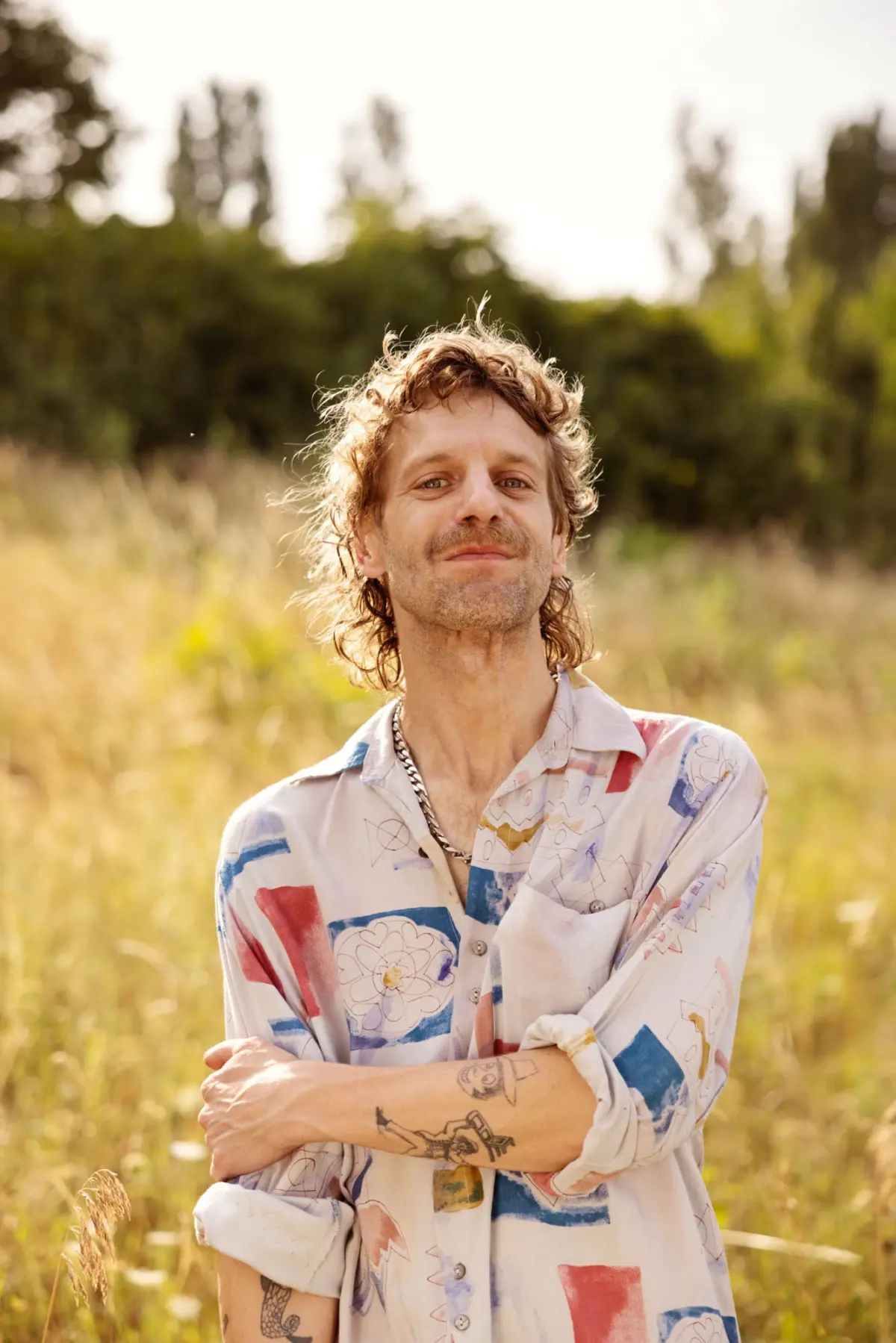 Person with wavy brown hair and tattoos stands in a sunlit field, arms crossed, wearing a colorful abstract-patterned shirt.