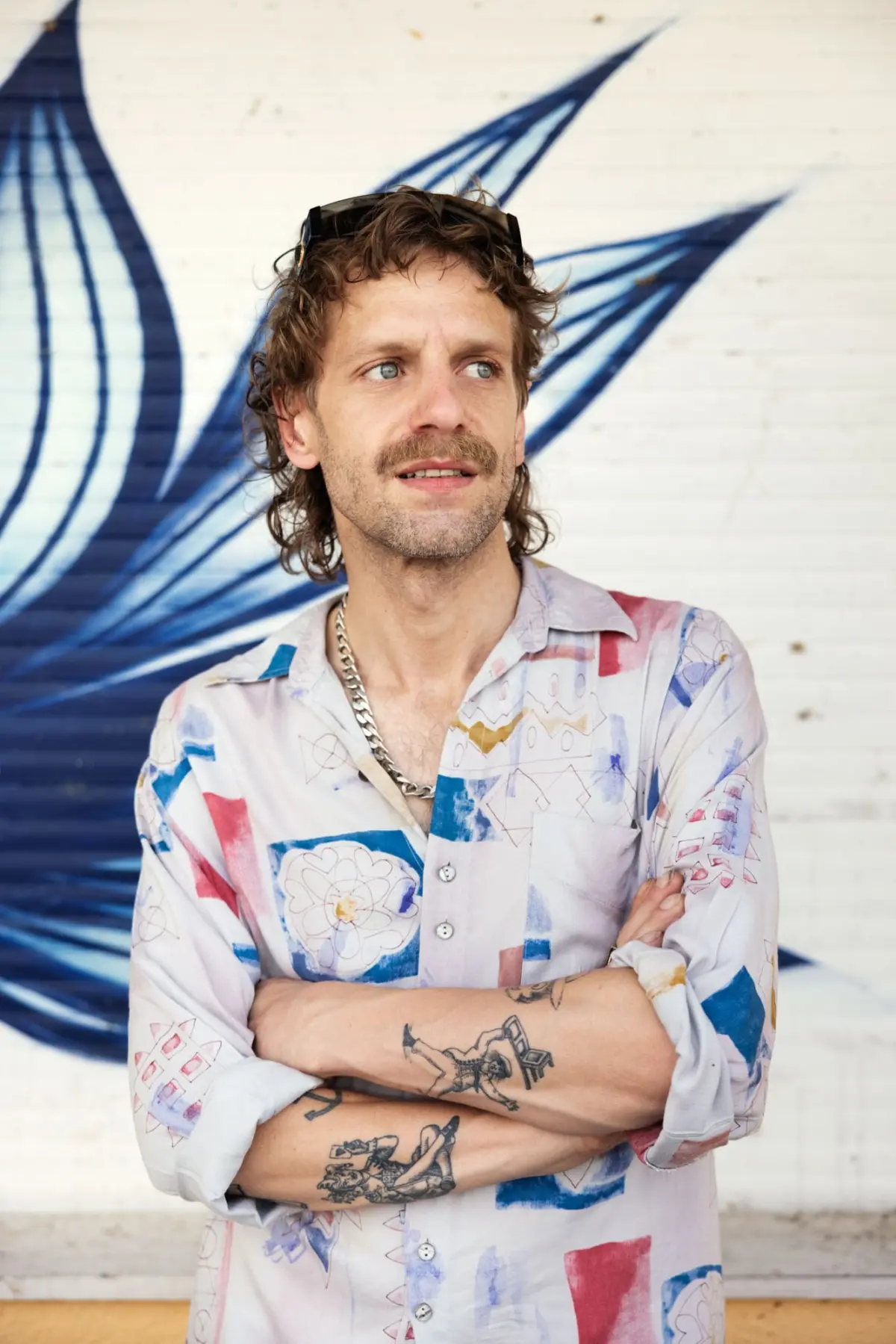 Adult male with tattoos and wavy brown hair, wearing a patterned shirt and sunglasses perched on his head, arms crossed before a blue graffiti mural.