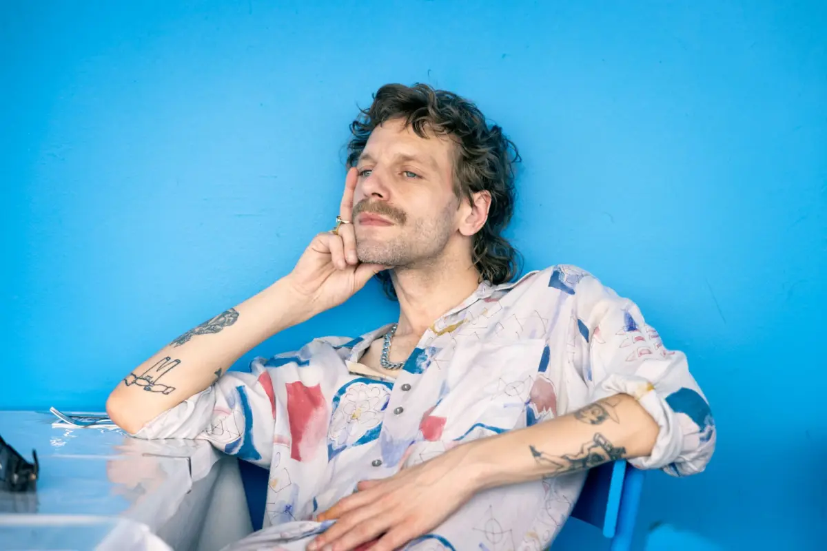 Tattooed adult with mustache rests chin on hand against a bright blue wall, seated at a counter.