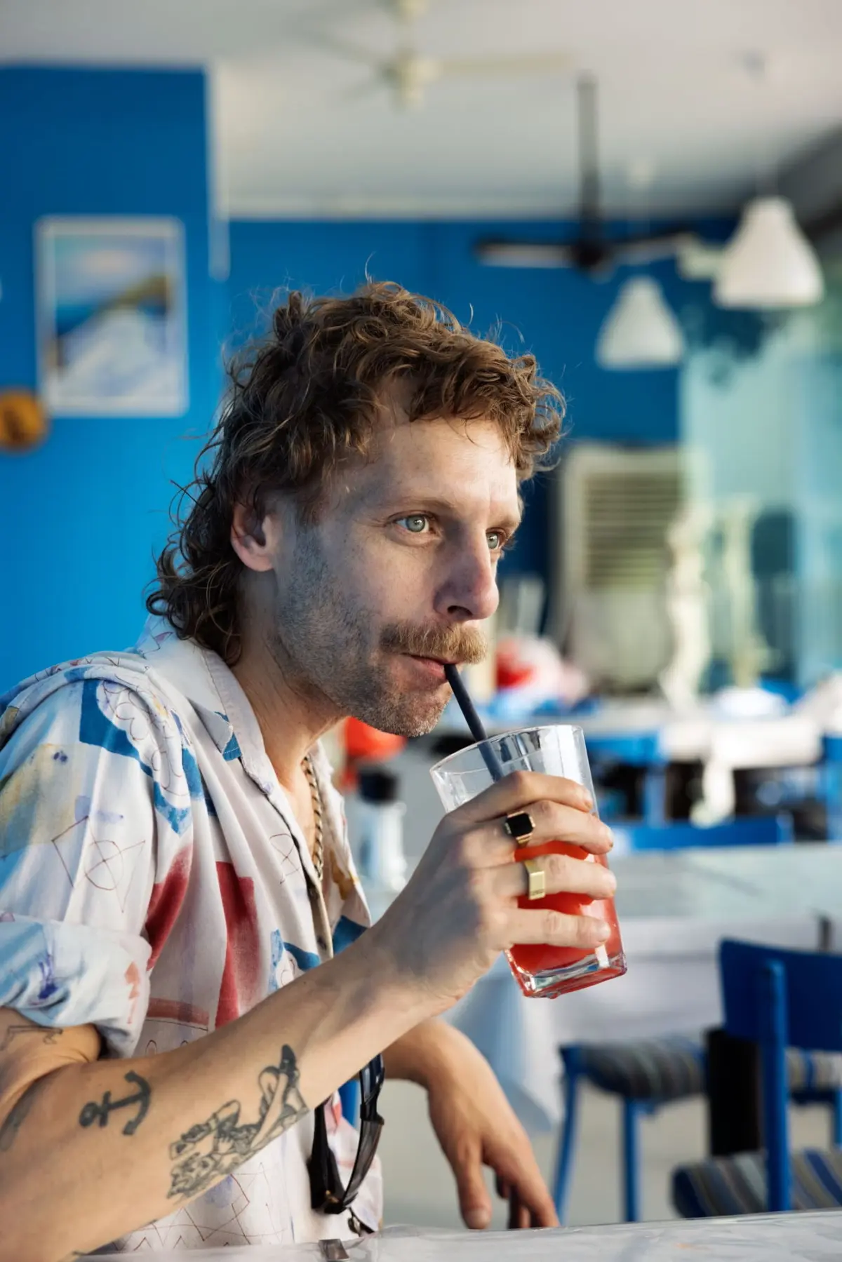 Adult male with curly hair drinks a red beverage through a straw at a blue-walled cafe.