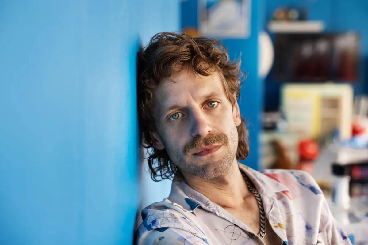 Adult person with mustache leaning against a blue wall indoors, with blurred shelves in background.