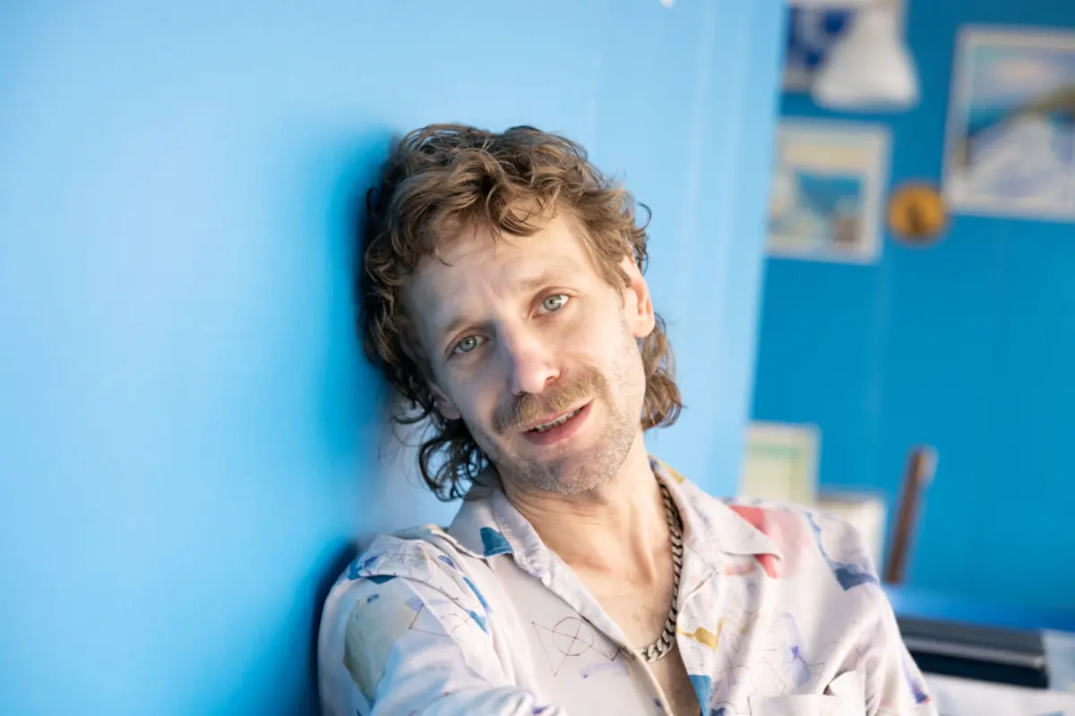 Man with wavy brown hair and stubble leaning against a blue wall in a bright room with framed pictures.