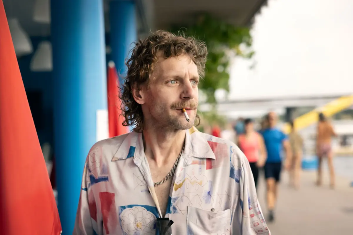 Adult man with cigarette in mouth, looking to the side, standing beside colorful poles on a beachside boardwalk.