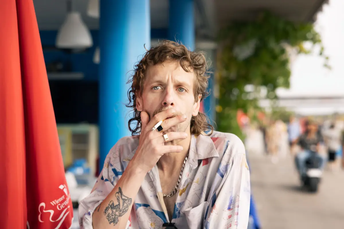 Adult man with tattoos smoking a cigarette outdoors beside blue columns, blurred crowd in the background.