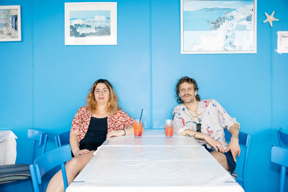 Two people sit at a marble-top table in a bright blue-walled cafe, with colorful drinks; framed artwork on the wall behind.