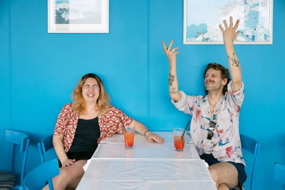 Two people at a bright blue cafe; one laughs with an orange beverage, the other raises arms in celebration.