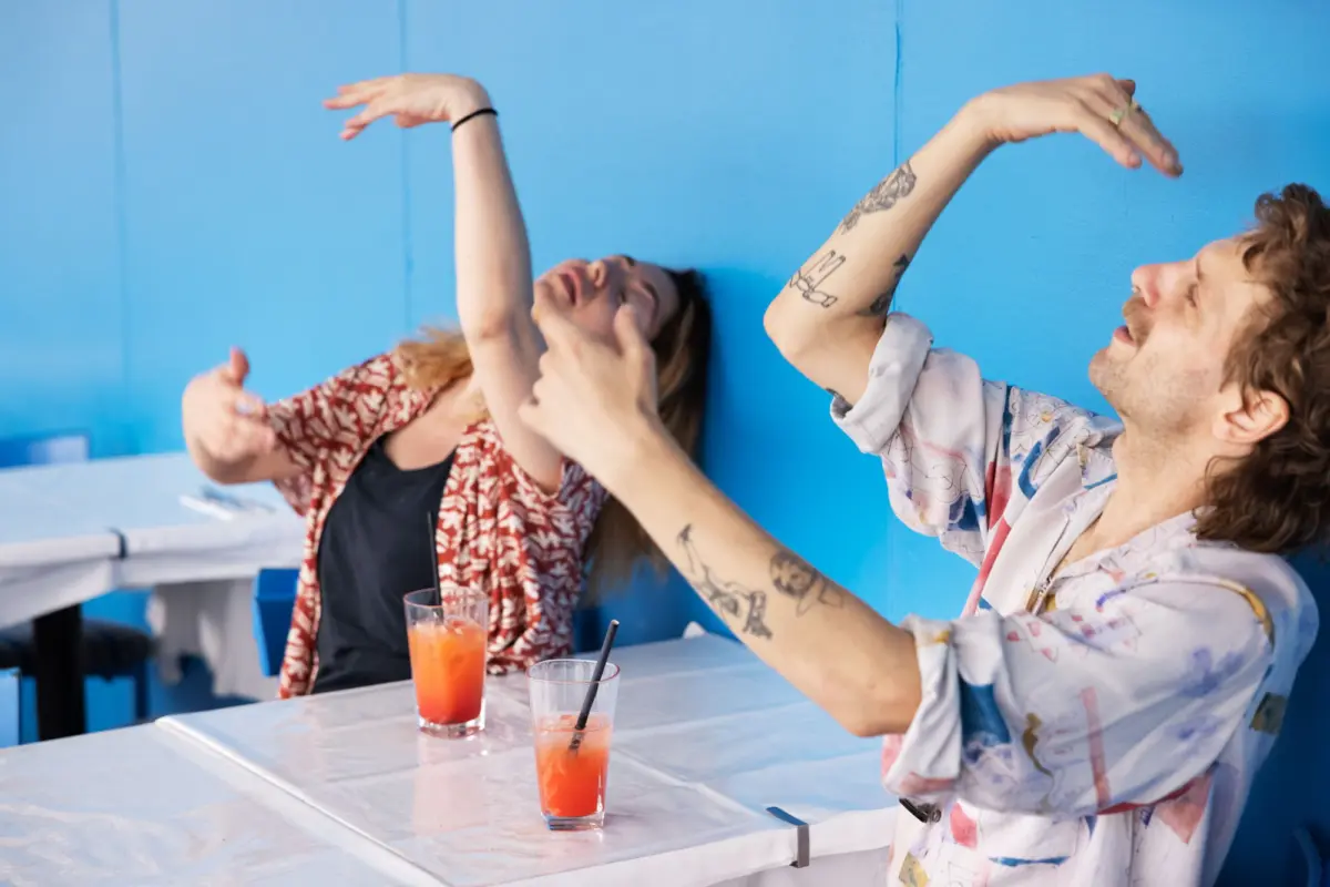 Two people celebrate with arms raised beside a bright blue wall; orange cocktails on a foil-covered table.