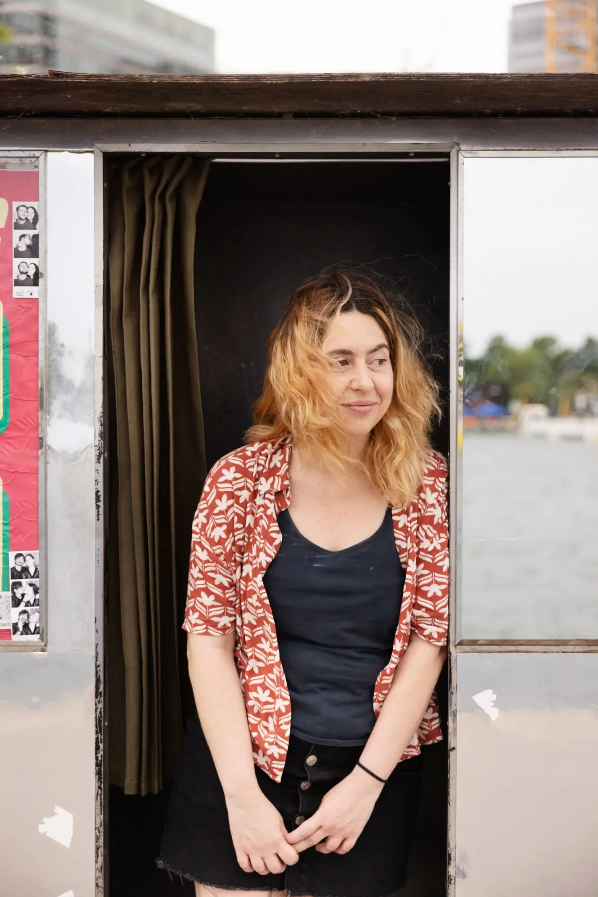 Person standing in doorway of a small metallic booth by the water, wearing a red floral shirt over a black top, looking to the side.