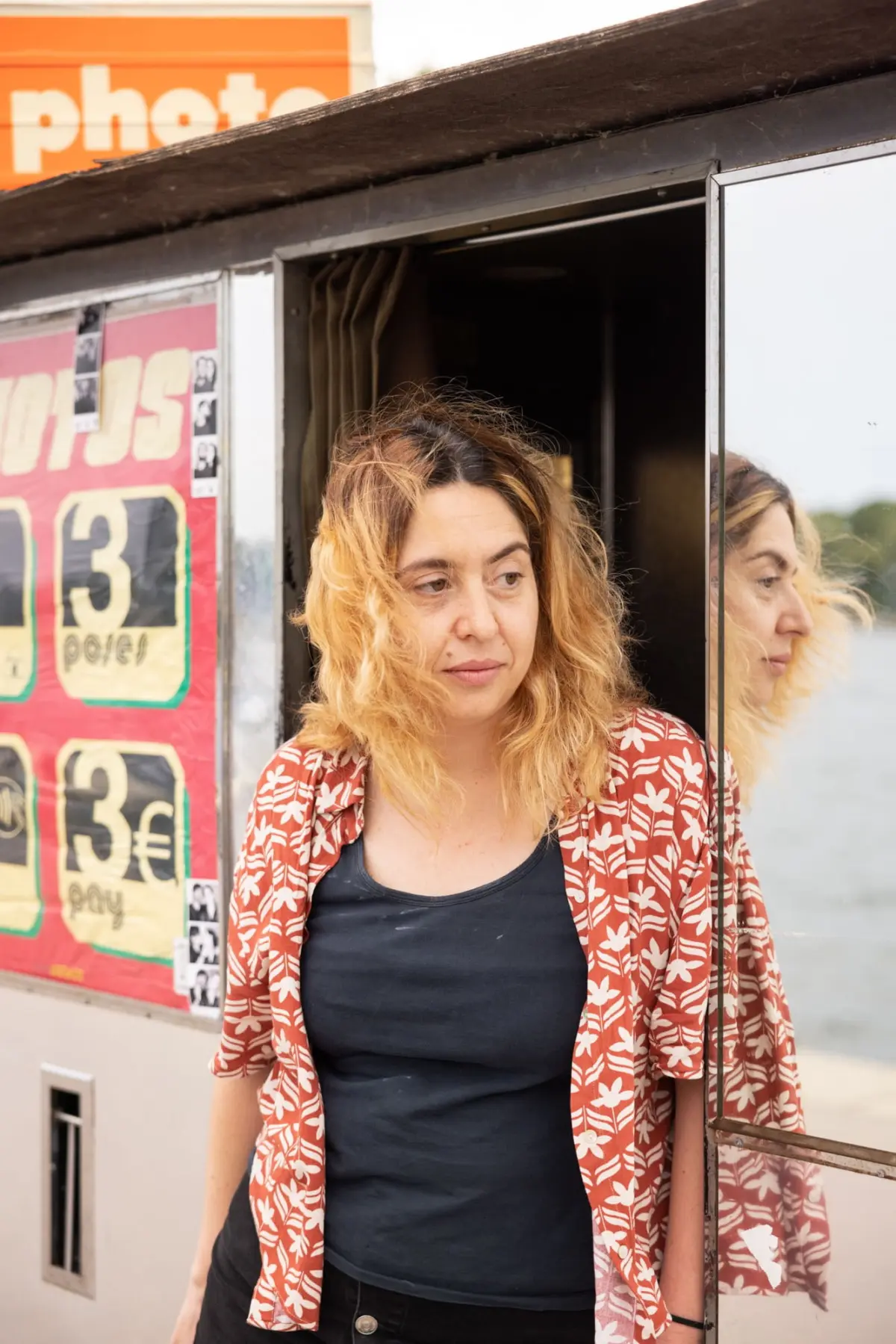 Person with blond hair wearing a red floral shirt over a dark top stands beside a trailer, looking to the side.