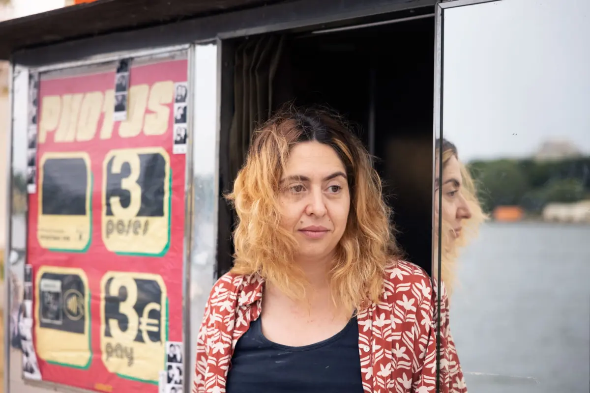 Person with wavy blonde hair stands beside a glass storefront, wearing a red floral shirt over a dark top, looking to the side.