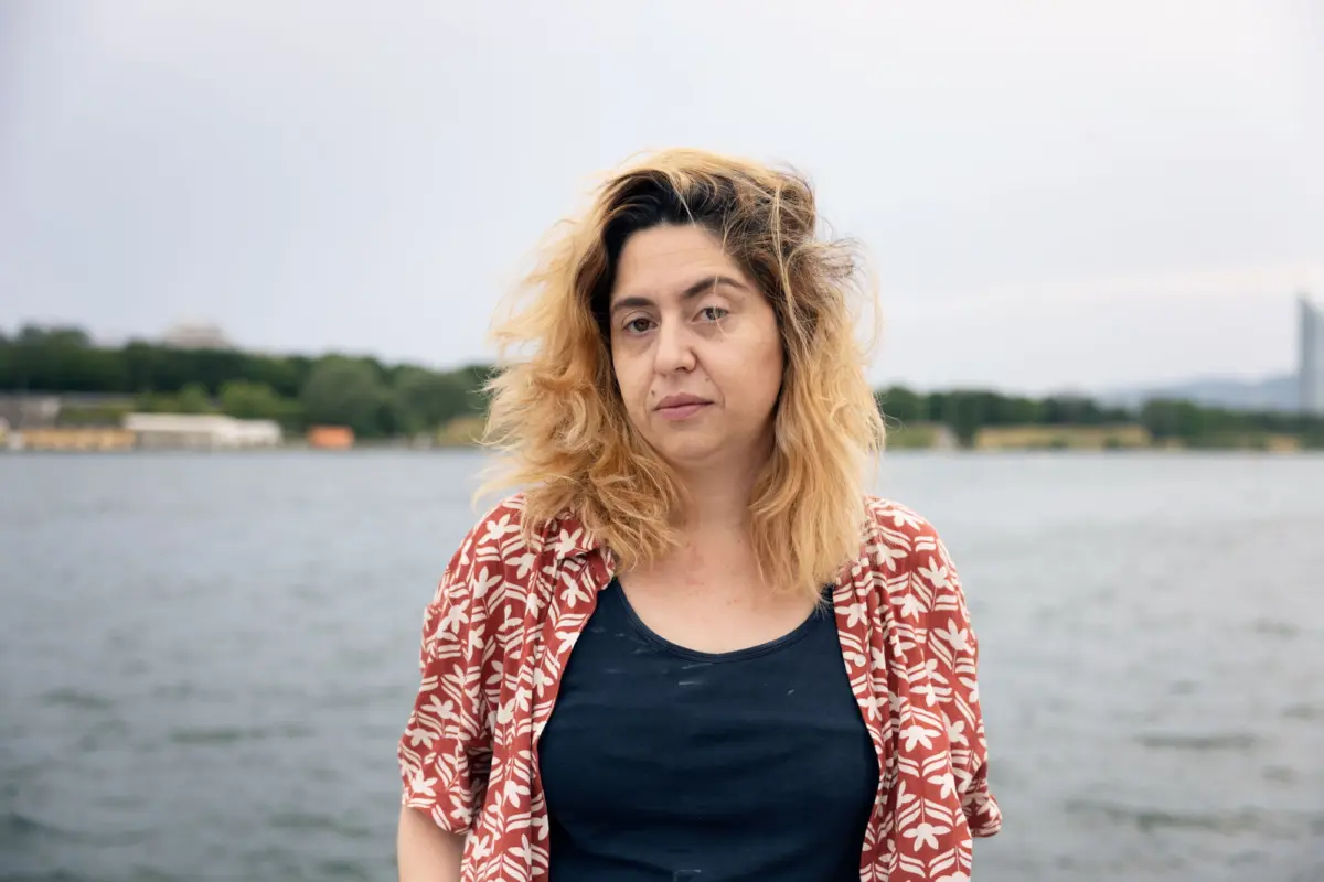 Person with wavy blonde hair stands by a lake, wearing a red floral-pattern shirt over a navy top.