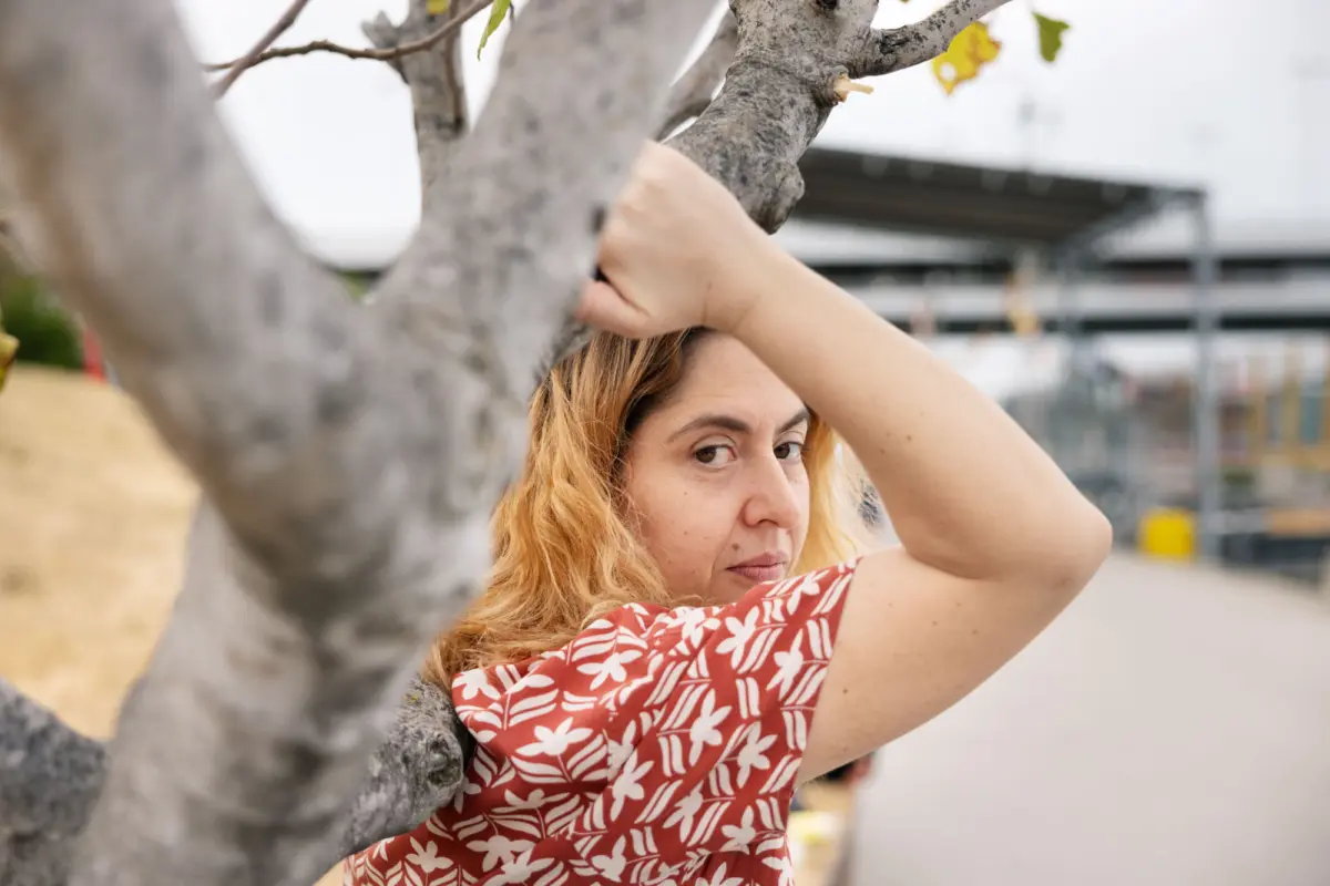 Person with red hair grips a tree trunk and peeks out in an outdoor urban setting.
