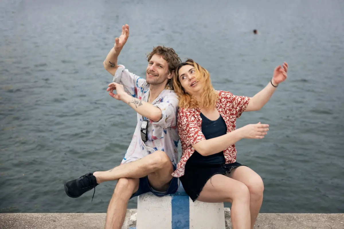 Two adults sit on a blue and white striped post by the water, arms raised in a playful pose.