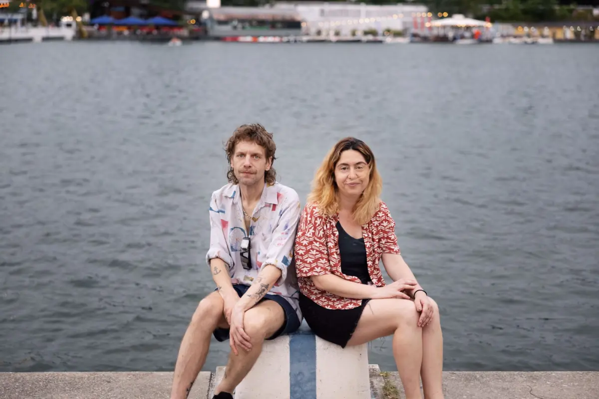 Two adults sit together on a concrete barrier by a harbor, with water and distant boats in the background.