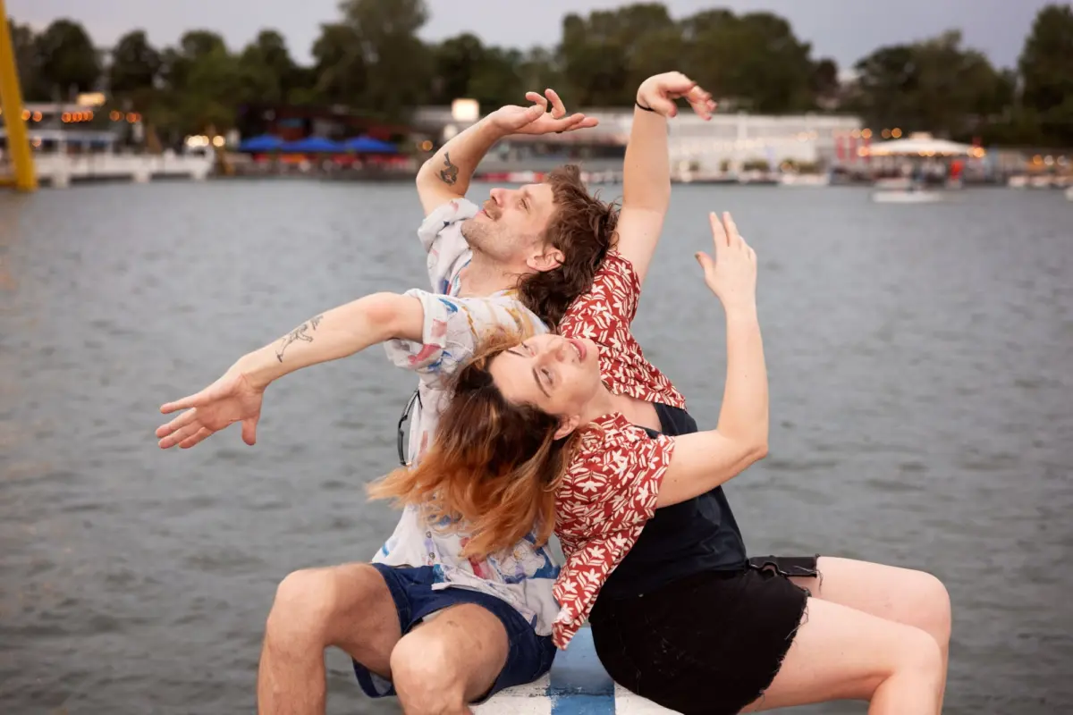 Two adults perform an acrobatic lift on a buoy by a lake, with a blurred shoreline and string lights in the background.