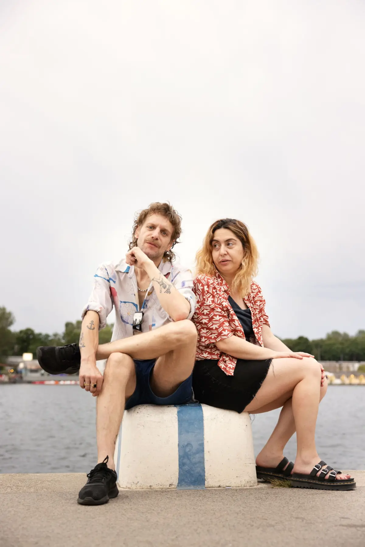 Two adults sit on a white and blue striped buoy by a waterfront, relaxing and looking toward each other as cloudy skies loom.