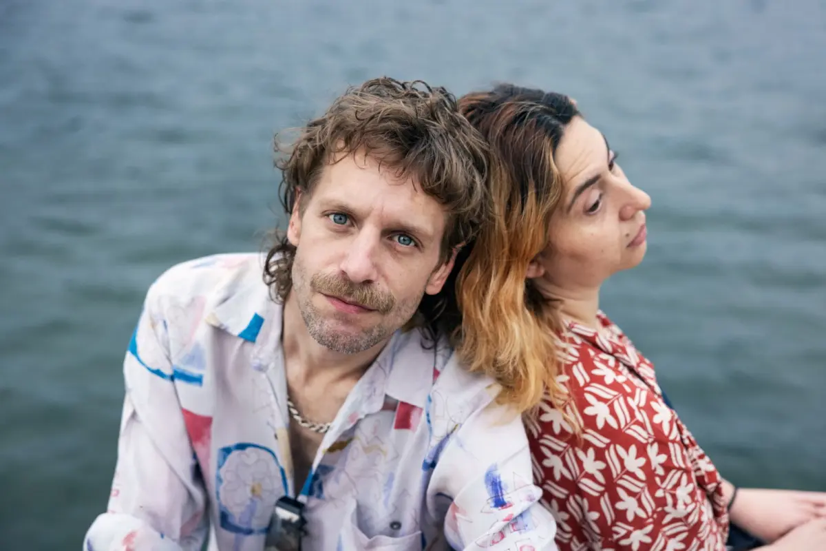 Adult with blue eyes and stubble in a colorful patterned shirt leans toward the camera; another adult with long hair beside them, by the sea.