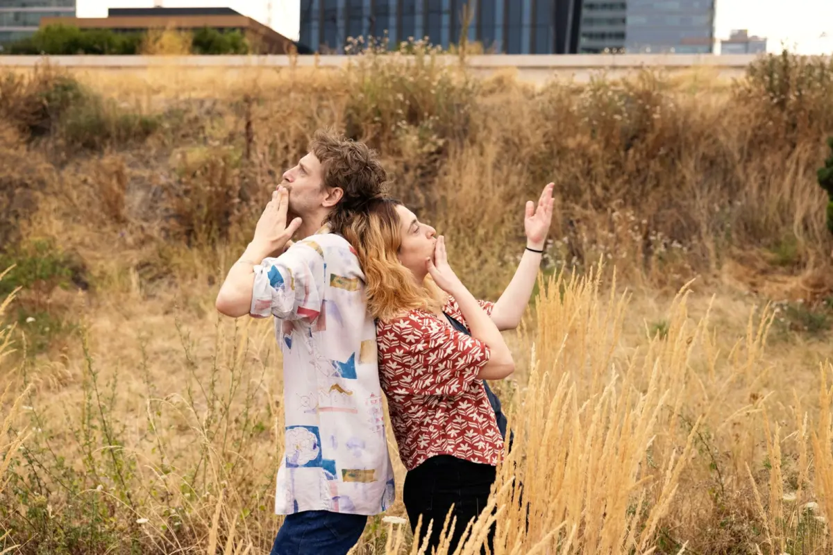 Two people stand back-to-back in a dry tall-grass field, gesturing toward their mouths; urban buildings visible behind.
