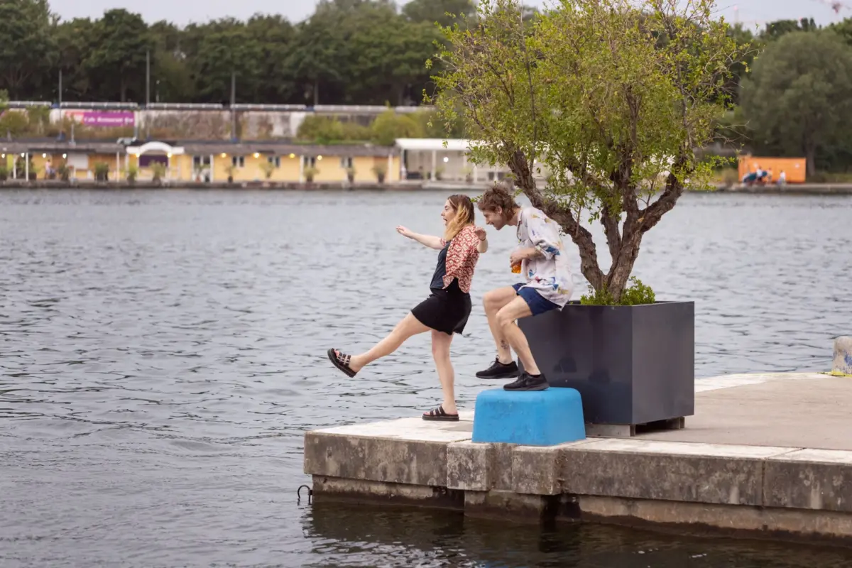 Two people jump from a concrete platform into a lake, beside a planter with a tree; urban waterfront buildings in the background.