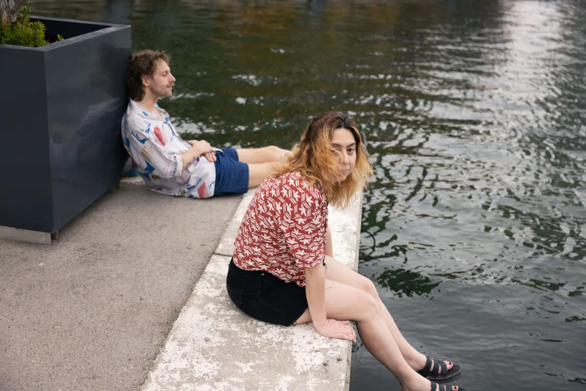 Two people by a riverside; one leans on a dark planter, the other sits on a concrete ledge in a red floral blouse.