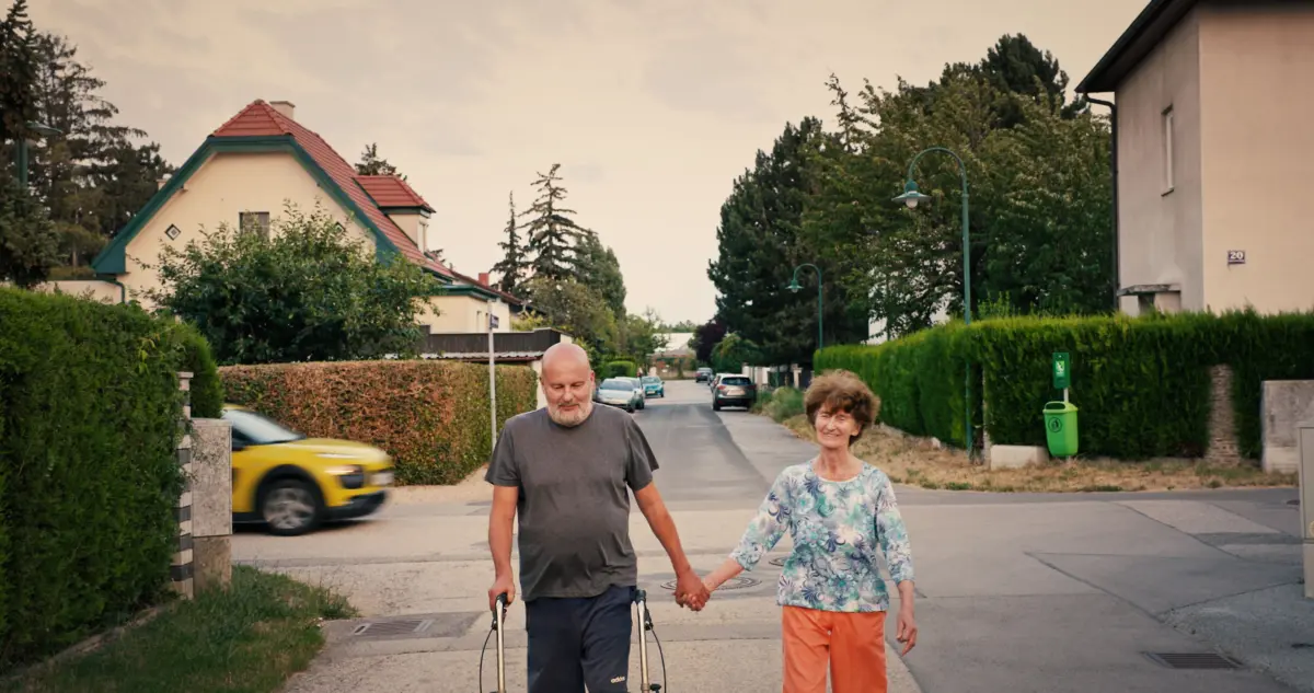 Elderly couple walking hand-in-hand down a suburban street; one uses a walker, the other smiles beside.