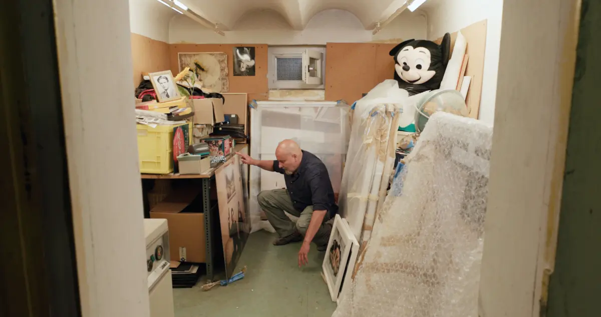 Bald person crouches beside cluttered storage room shelves, sorting items among framed photos, boxes, bubble wrap, and a large Mickey Mouse head prop.