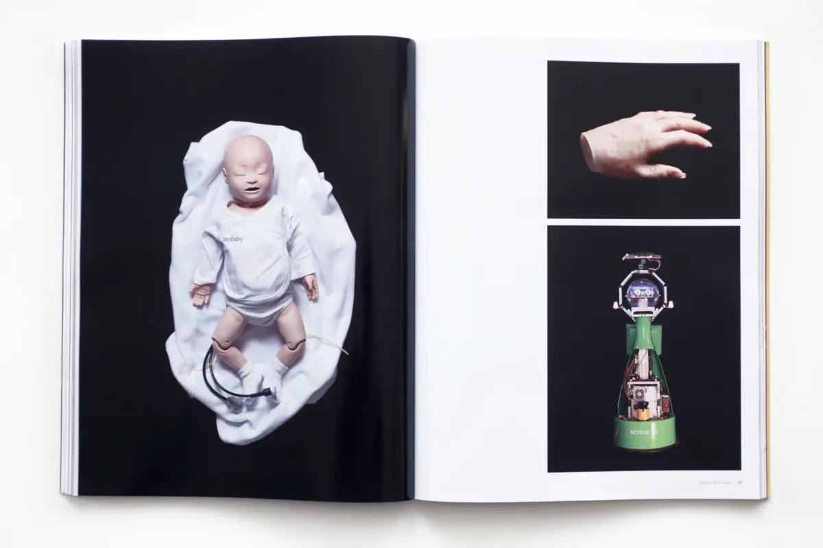 Open book spread: left page shows a baby-doll lying on white cloth; right page shows a pale hand on black background above a green robotic device.