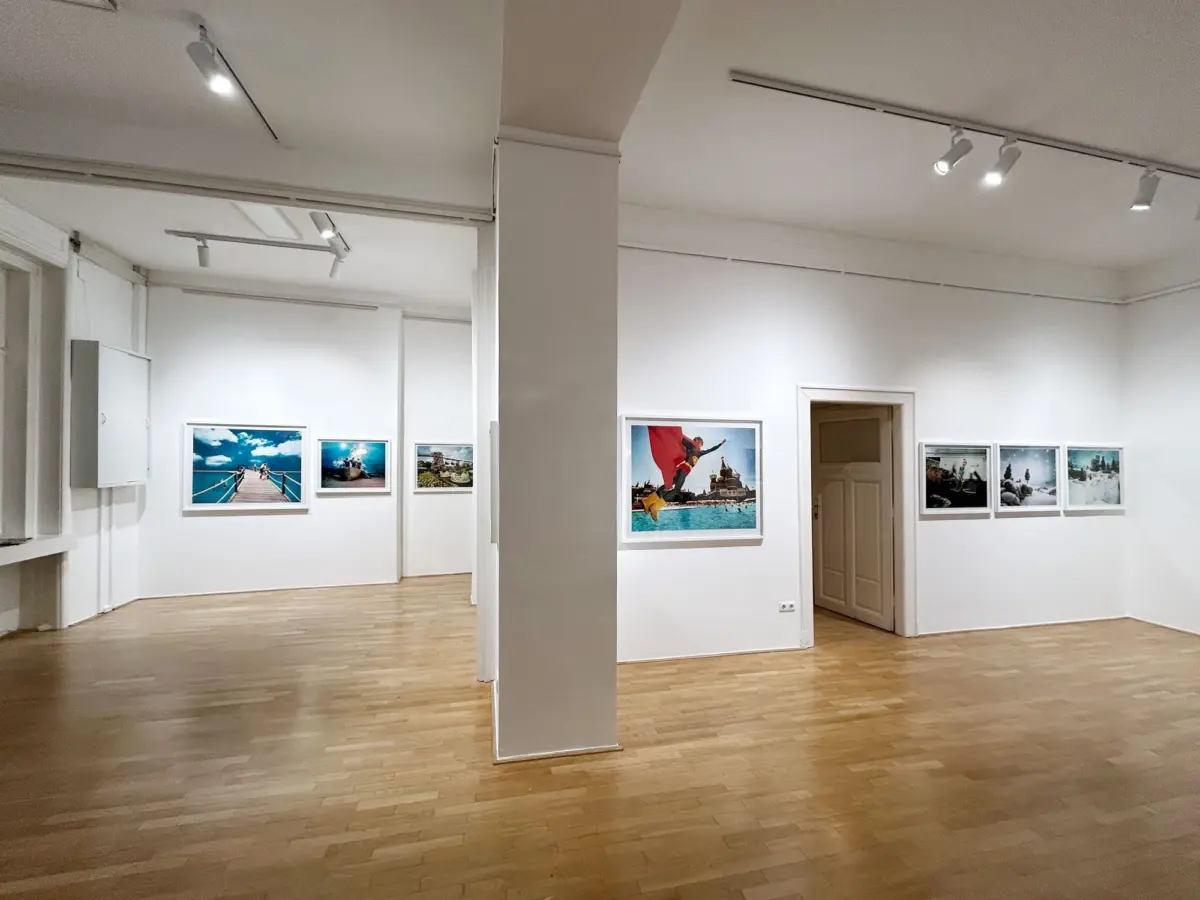 Interior of a white-walled art gallery with a wooden floor; framed photographs along the walls and a central column under track lighting.