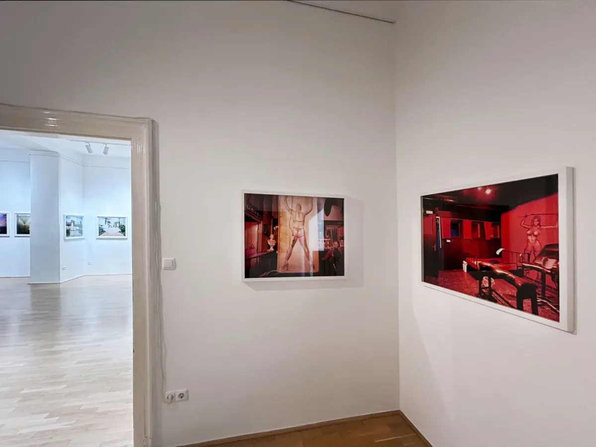Art gallery interior with white walls; two framed photographs on the right and a doorway to a brighter room with more artwork.