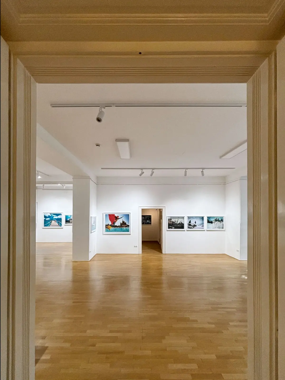 Interior of a white-walled art gallery viewed through a doorway; framed photographs hung along the walls, hardwood floor.