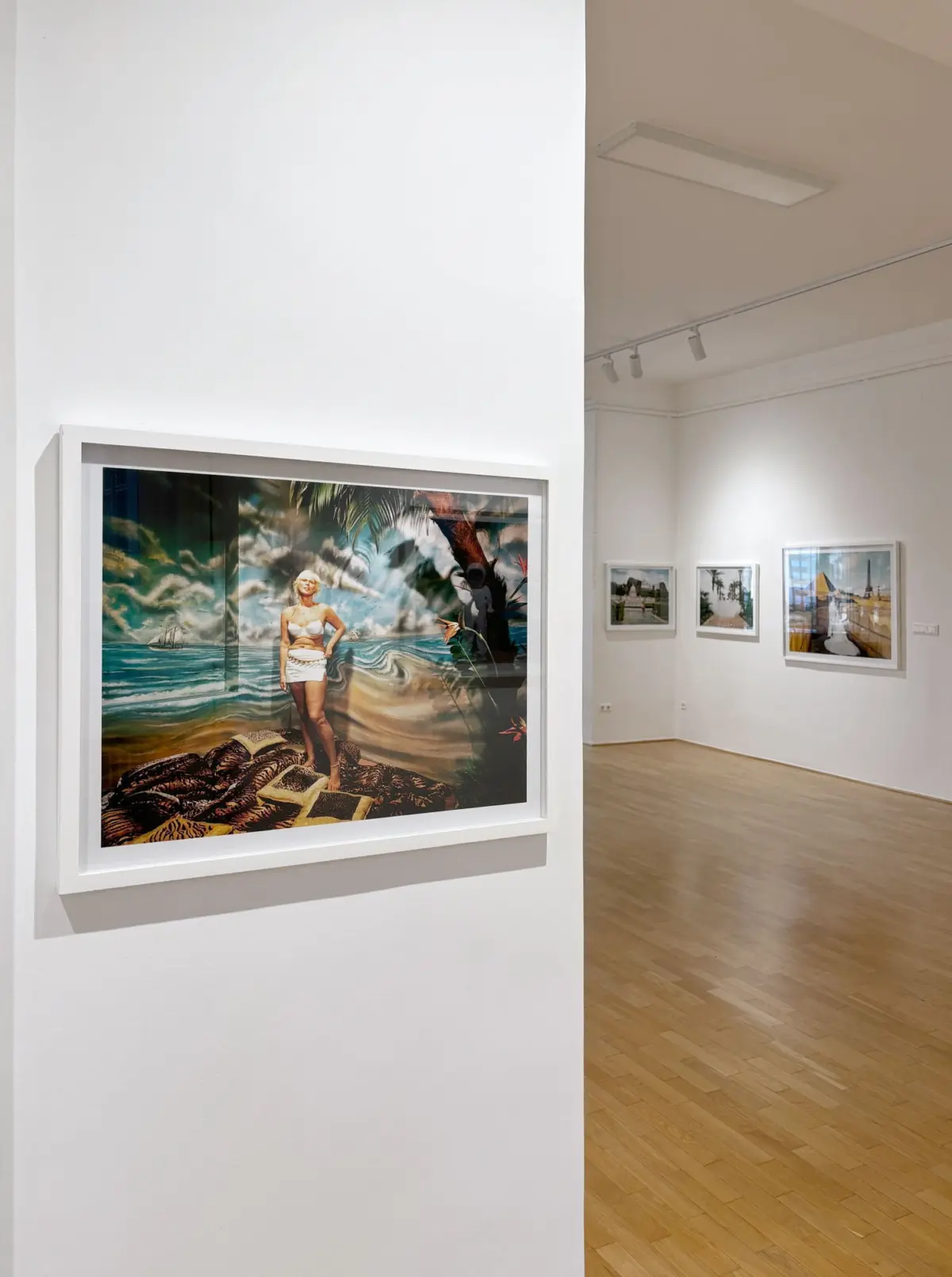 Framed photograph of a person standing on a rocky tropical beach with palm trees and ocean, displayed in an art gallery.