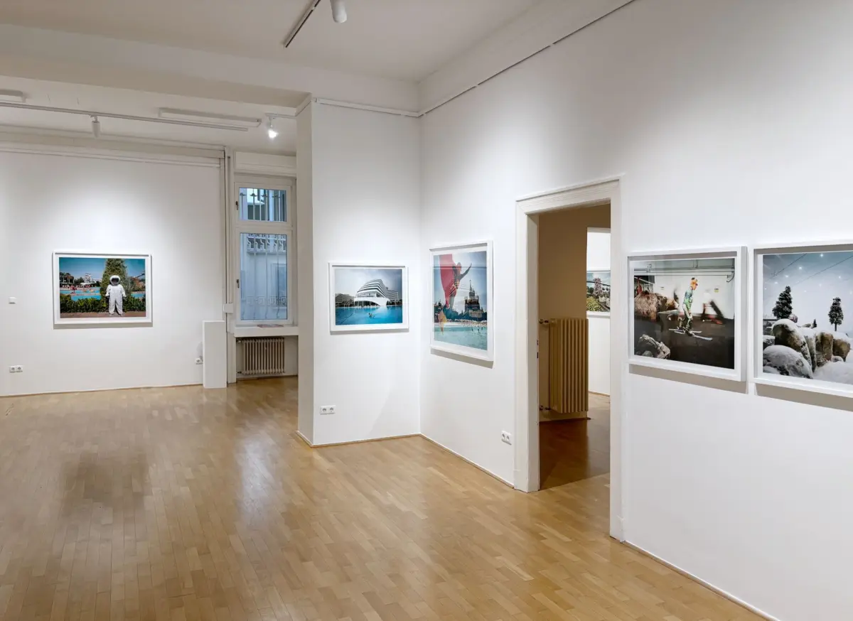 Art gallery interior with white walls and wooden floor; framed photographs line the walls; doorway to another room; large window on the left.