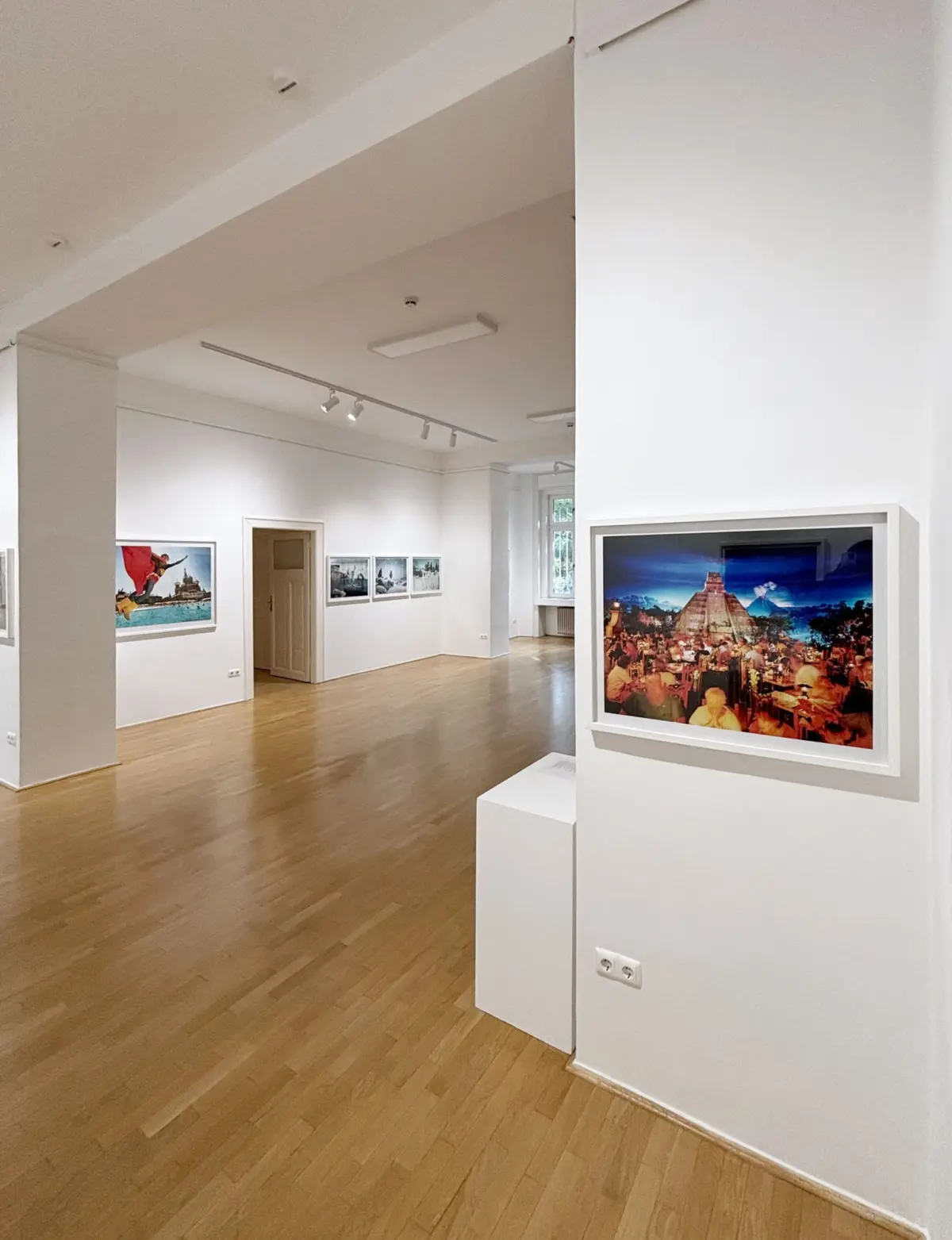 White-walled art gallery interior with wooden floor; framed photographs on walls, including a large colorful photograph.