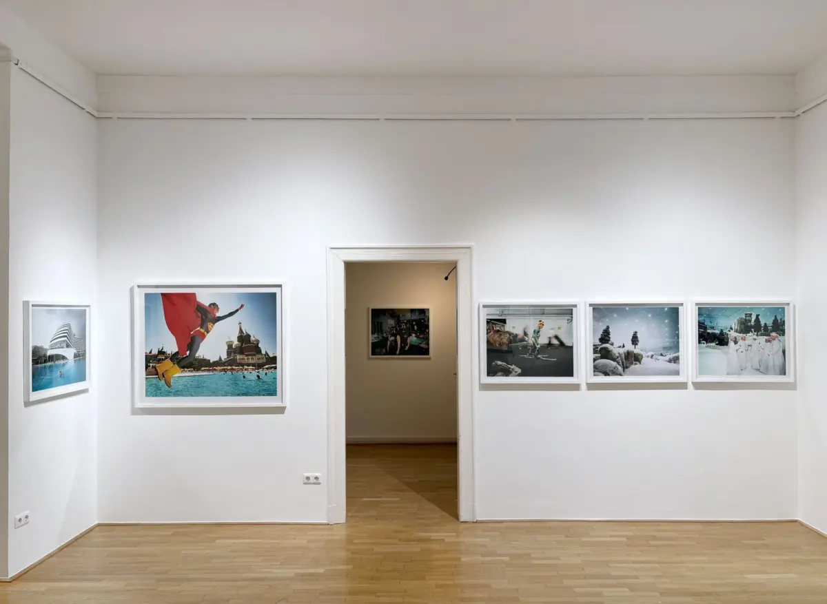 White-walled art gallery with framed photographs along the walls and a doorway in the center; light wooden floor.