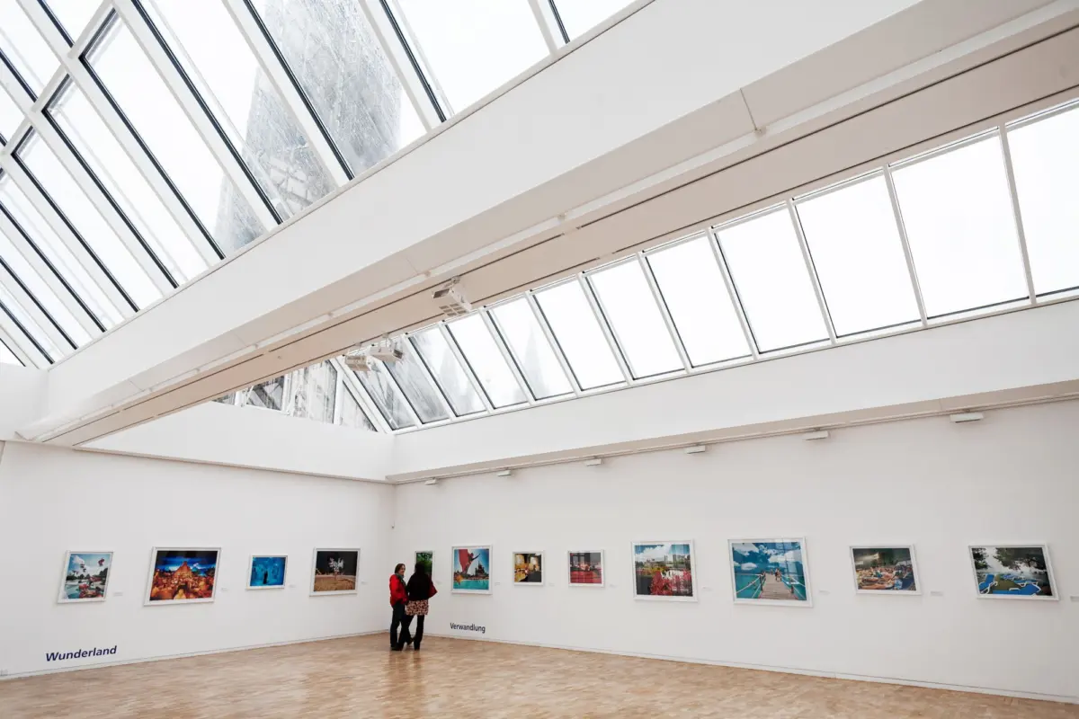 Two visitors examine framed photographs in a bright white art-gallery with a slanted glass skylight and wooden floor.