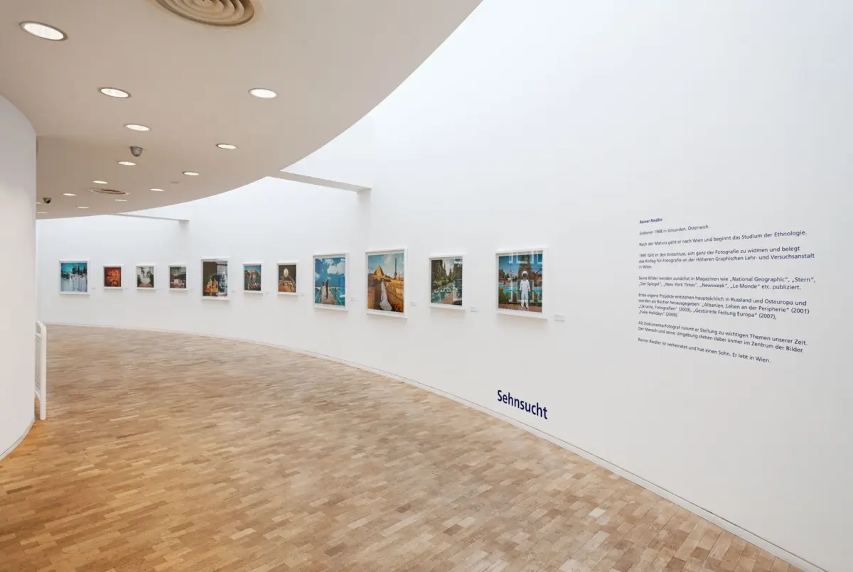 Curved white gallery interior with evenly spaced framed photographs along the wall; warm wooden floor and recessed ceiling lights.