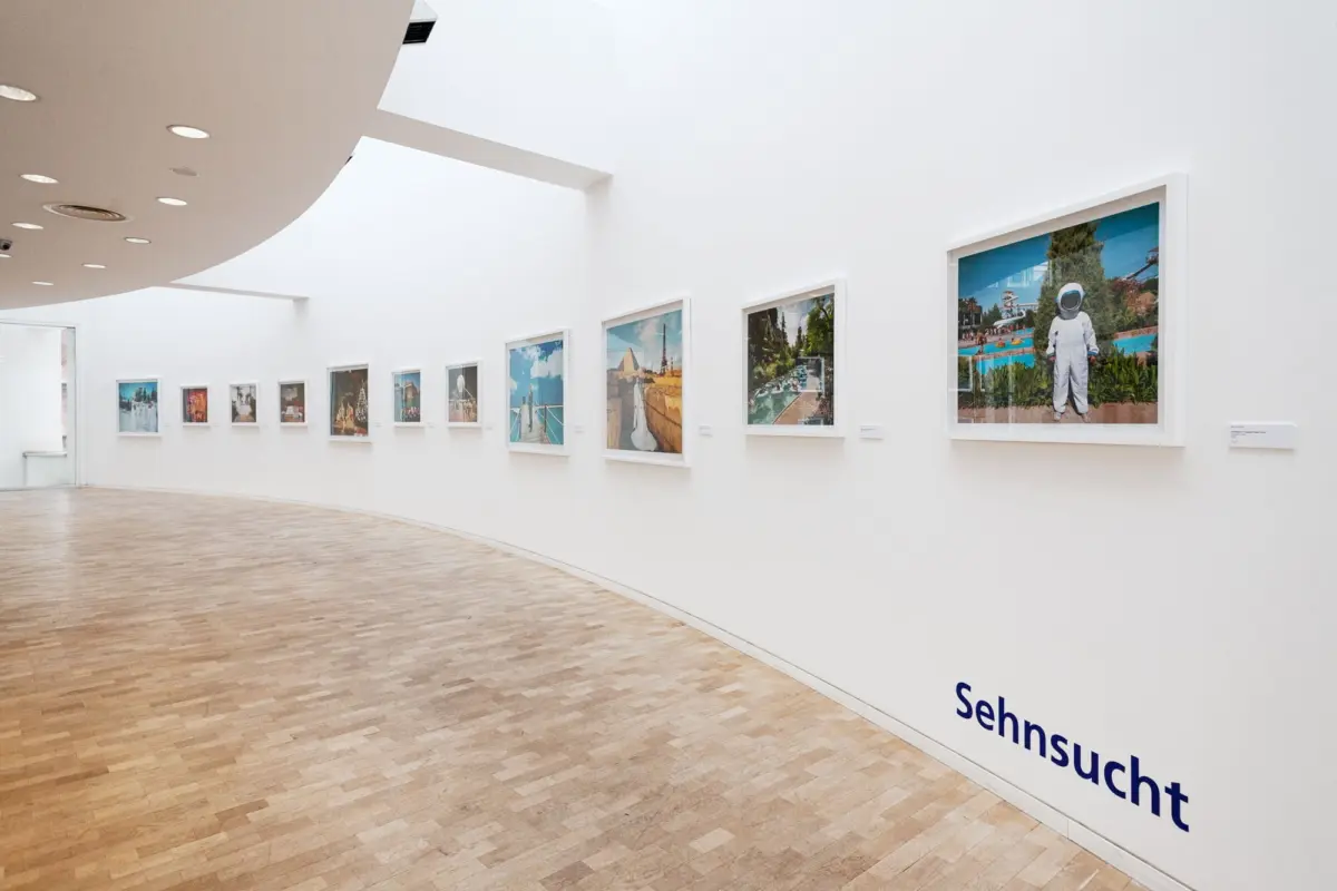 Bright white art gallery interior with a curved ceiling, a row of framed photographs along the wall, and a large astronaut image on the right; wooden floor.