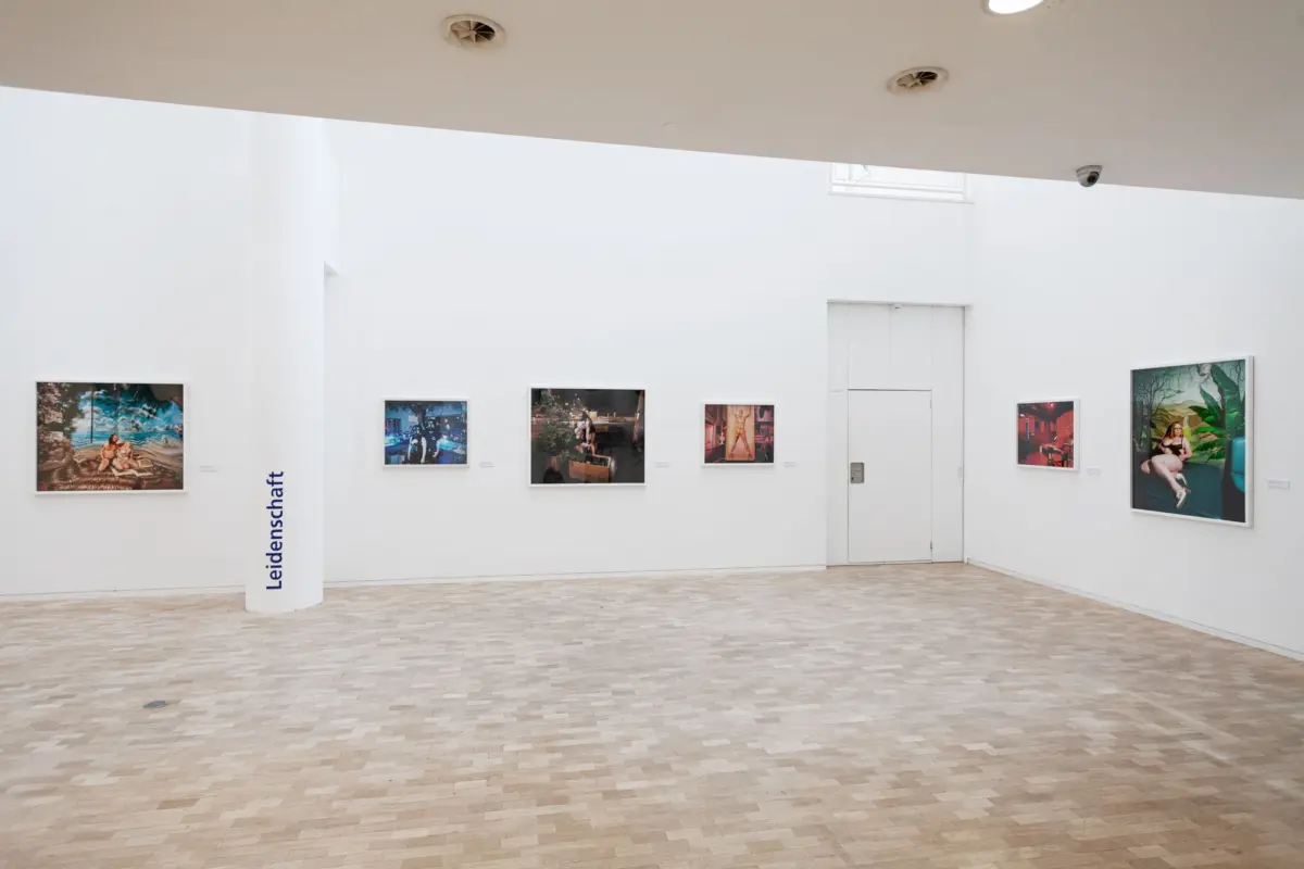 White-walled art gallery with framed photographs along the wall and a white pillar with blue text 'Leidenschaft'; light tiled floor.