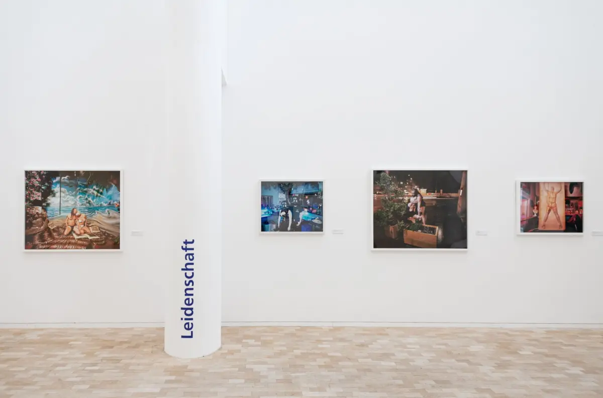 Four framed photographs hung on a white gallery wall with a white cylindrical column in the foreground and a beige tiled floor.