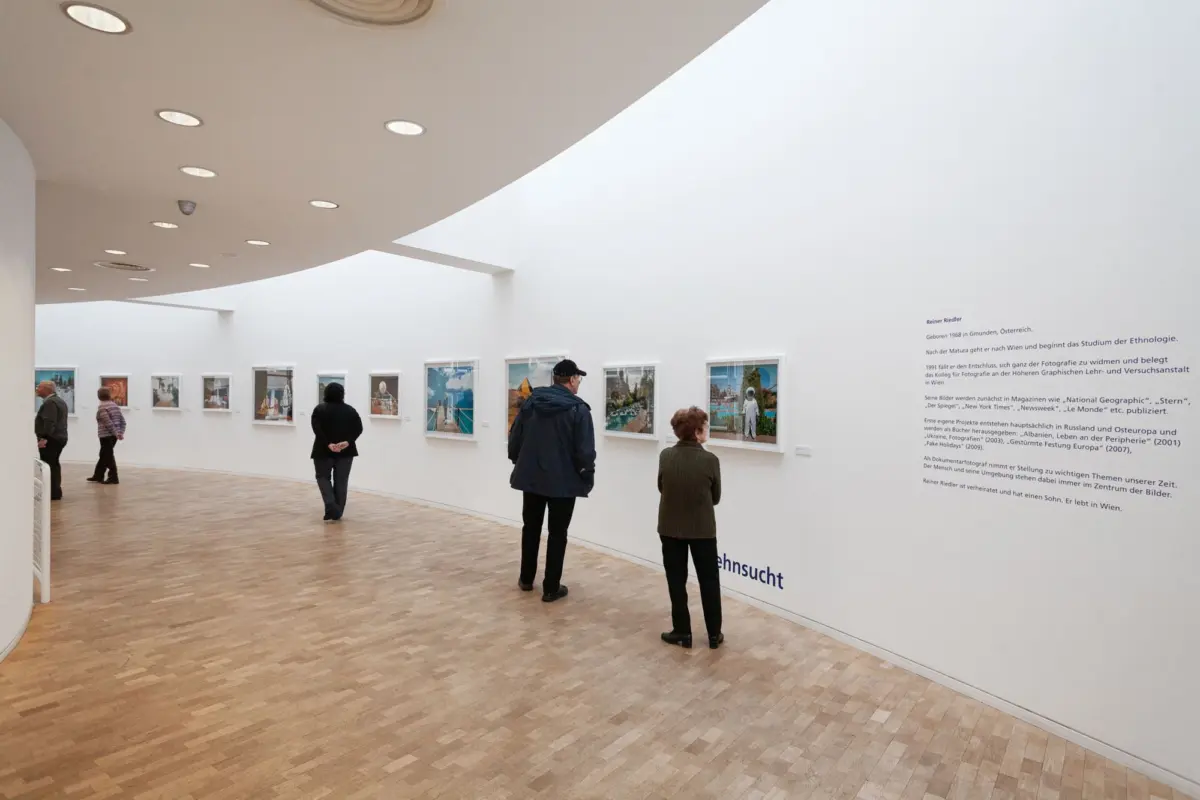 Gallery with white curved ceiling; visitors observe framed photographs along a curved wall; wooden floor, bright overhead lights.