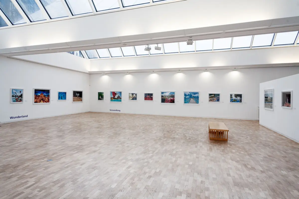 Spacious white-walled art gallery with a skylight roof, framed photographs along the walls, and a wooden bench in the foreground.