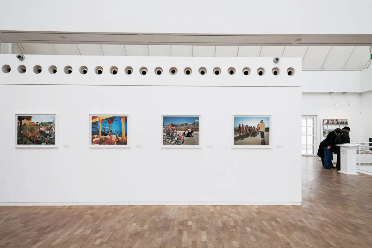 White-walled art gallery with a row of circular vents above four framed photos; visitors browse at right.